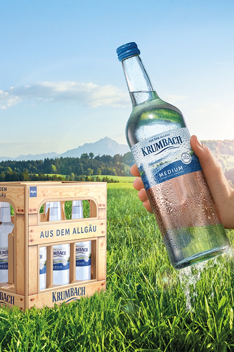Hand hält Krumbach Mineralwasser (Medium) vor Allgäu-Kulisse, daneben 6er-Kasten