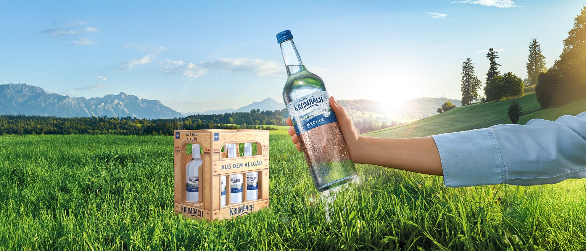 Hand hält Krumbach Mineralwasser (Medium) vor Allgäu-Kulisse, daneben 6er-Kasten