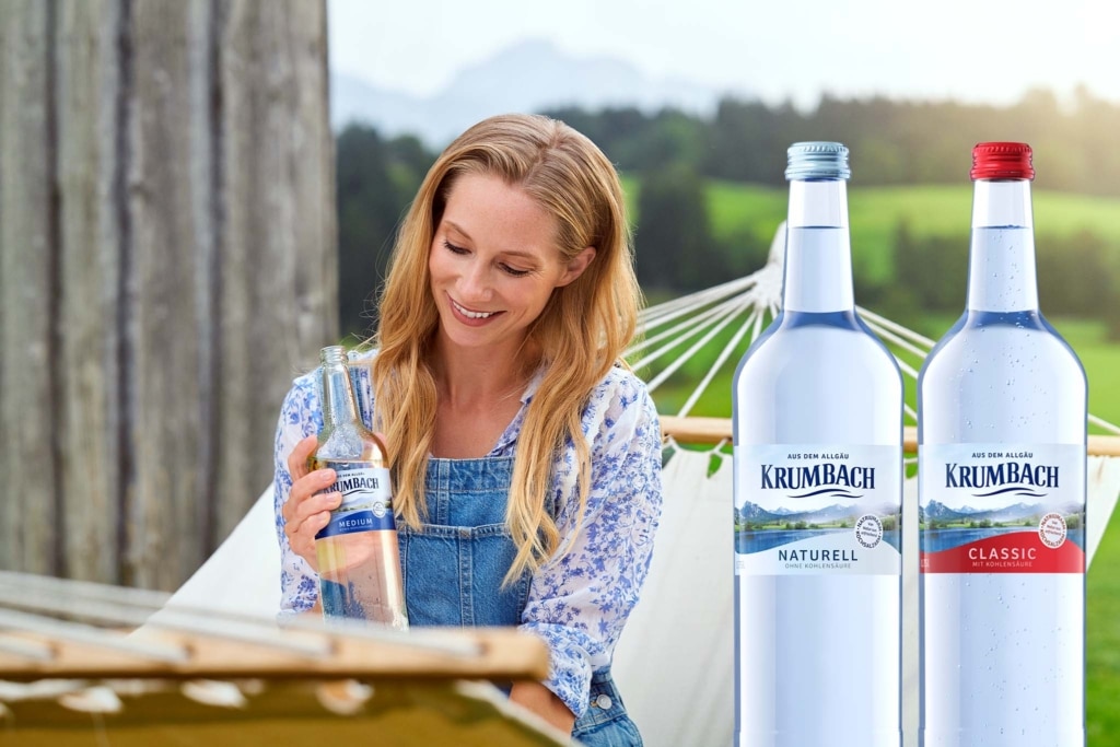 Frau sitzt in Hängematte mit Krumbach-Flasche in der Hand; daneben Flaschen Naturell und Classic vor Landschaft