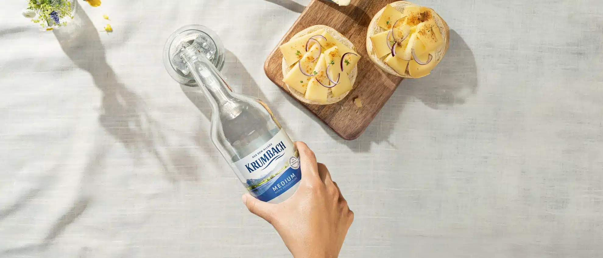 Hand gießt Krumbach Mineralwasser in ein Glas, daneben Käsebrote mit Käse und Zwiebeln.
