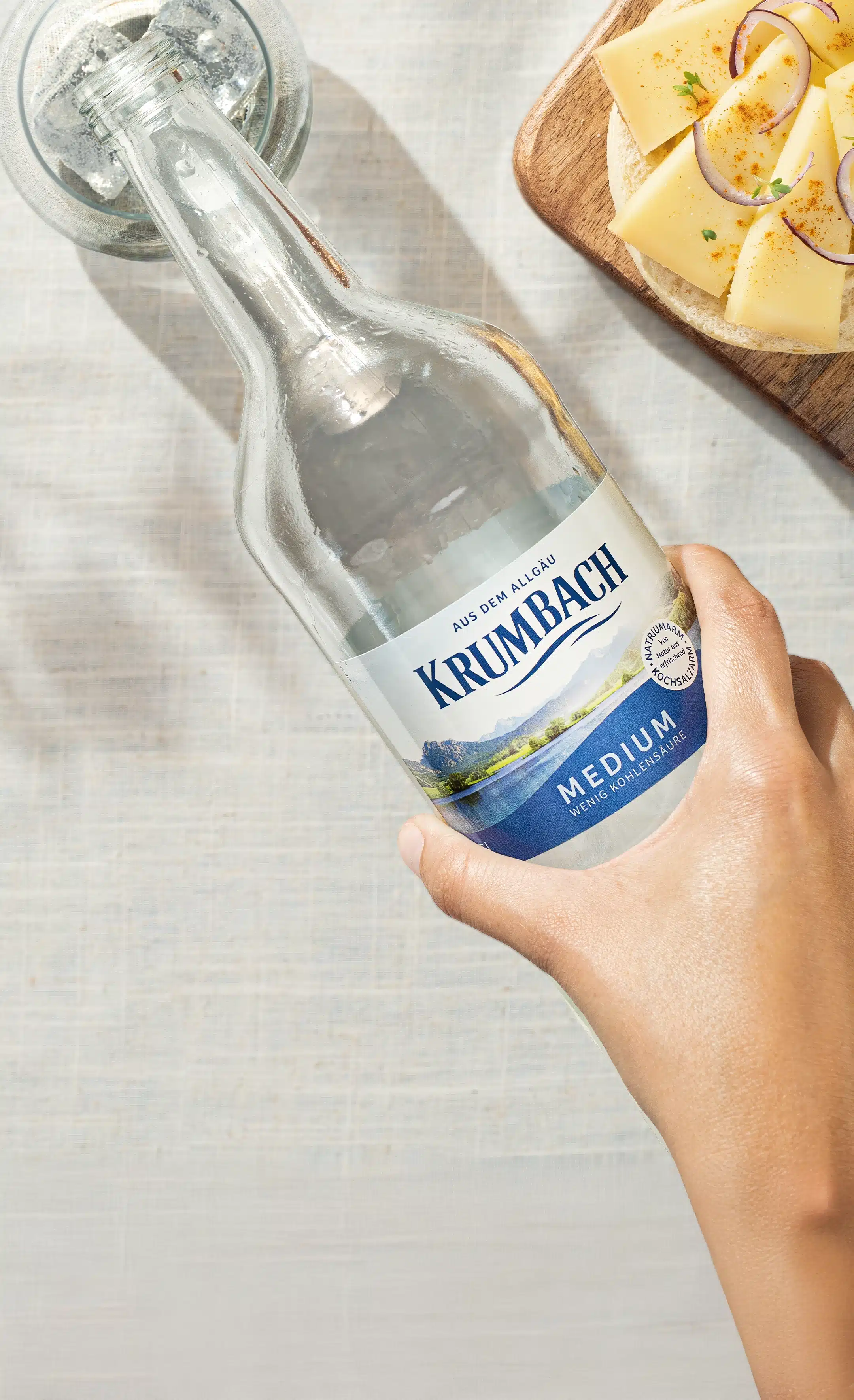 Hand gießt Krumbach Mineralwasser in ein Glas, daneben Käsebrote mit Käse und Zwiebeln.