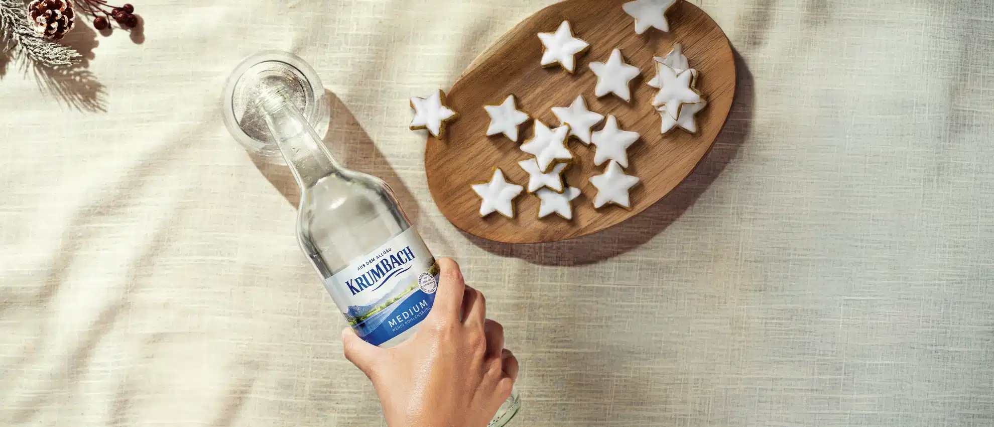 Hand gießt Krumbach Mineralwasser in ein Glas, daneben Holzschale mit Zimtsternen.