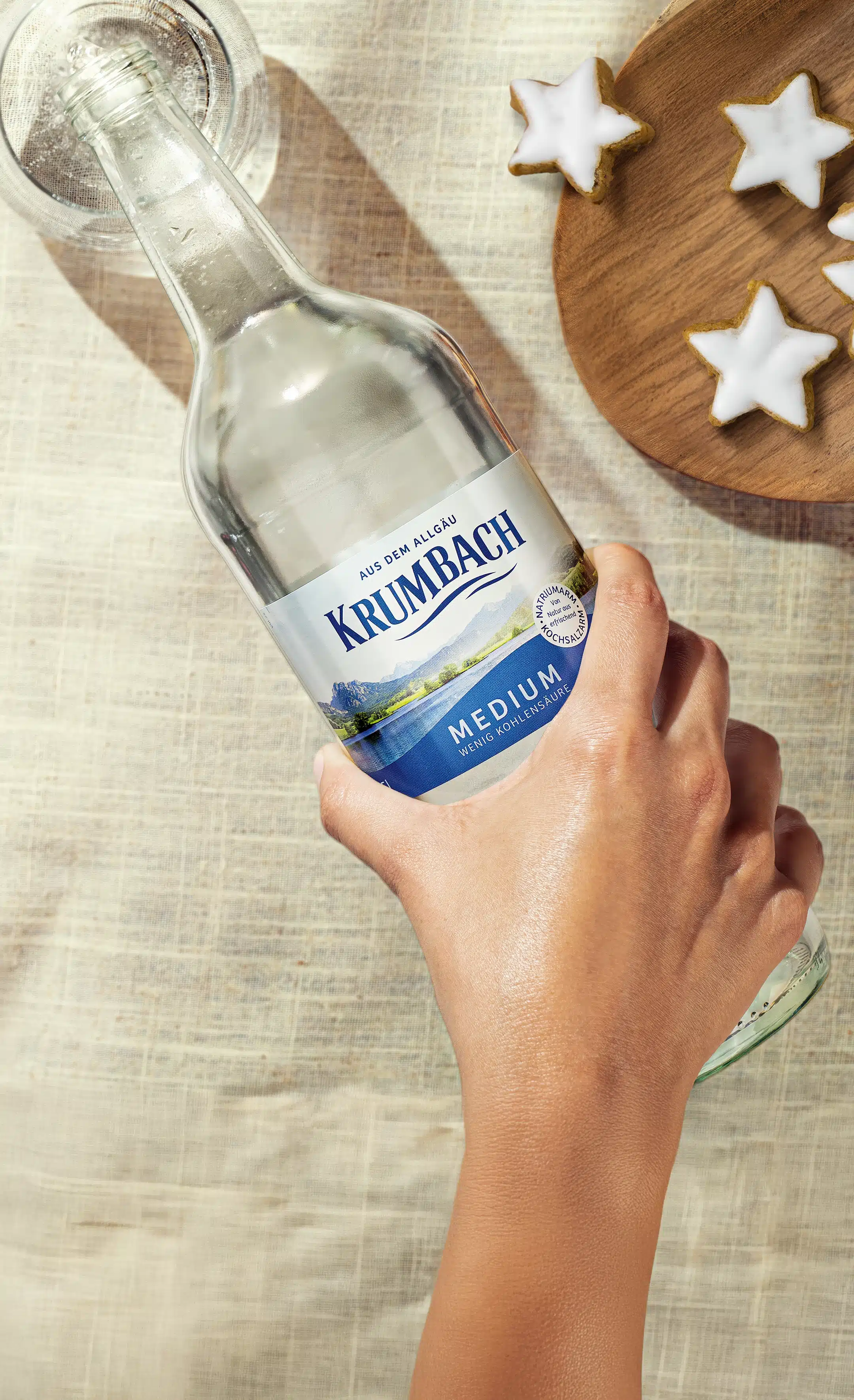 Hand gießt Krumbach Mineralwasser in ein Glas, daneben Holzschale mit Zimtsternen.