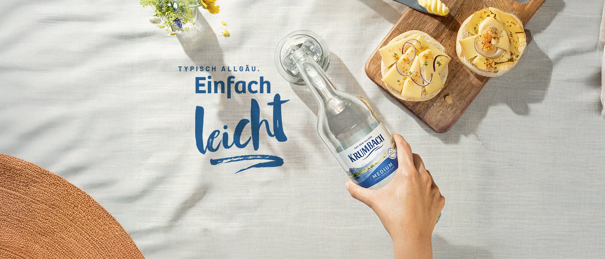 Hand hält eine Flasche Krumbach Mineralwasser auf gedecktem Tisch mit Brot und Käse, darüber Schriftzug ‚Typisch Allgäu. Einfach leicht.‘