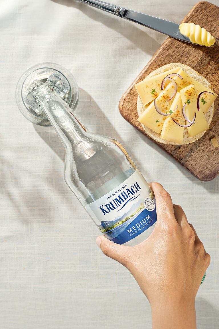 Hand hält eine Flasche Krumbach Mineralwasser auf gedecktem Tisch mit Brot und Käse, darüber Schriftzug ‚Typisch Allgäu. Einfach leicht.‘