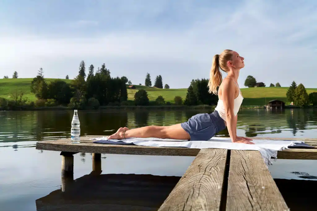 Frau macht Yoga auf einem Holzsteg am See, daneben steht eine Krumbach Naturell Glasflasche