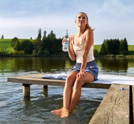 Frau sitzt mit Krumbach Naturell Glasflasche auf einem Holzsteg am See im Allgäu