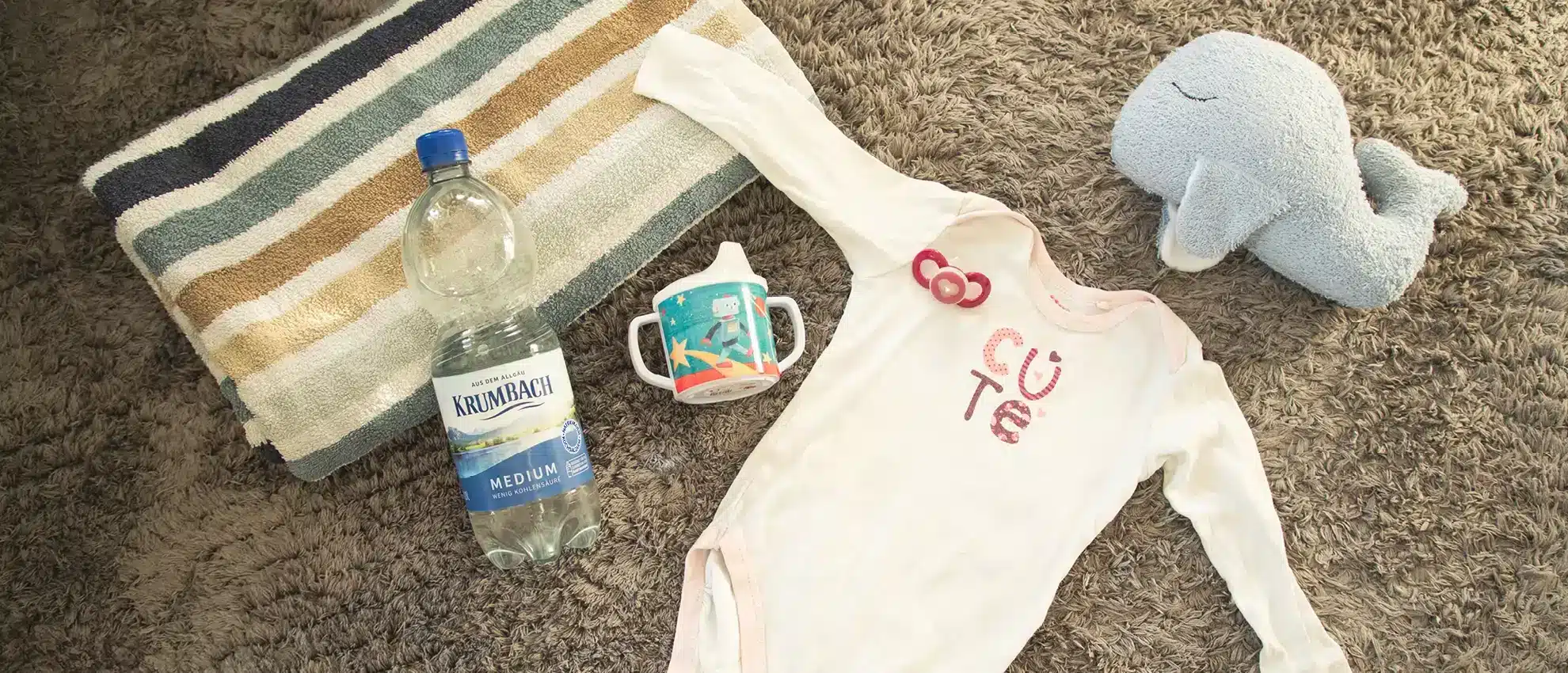 Krumbach Mineralwasser-Flasche neben Baby-Strampler und Trinkflasche
