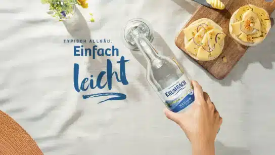 Hand hält eine Flasche Krumbach Mineralwasser auf gedecktem Tisch mit Brot und Käse, darüber Schriftzug ‚Typisch Allgäu. Einfach leicht.‘