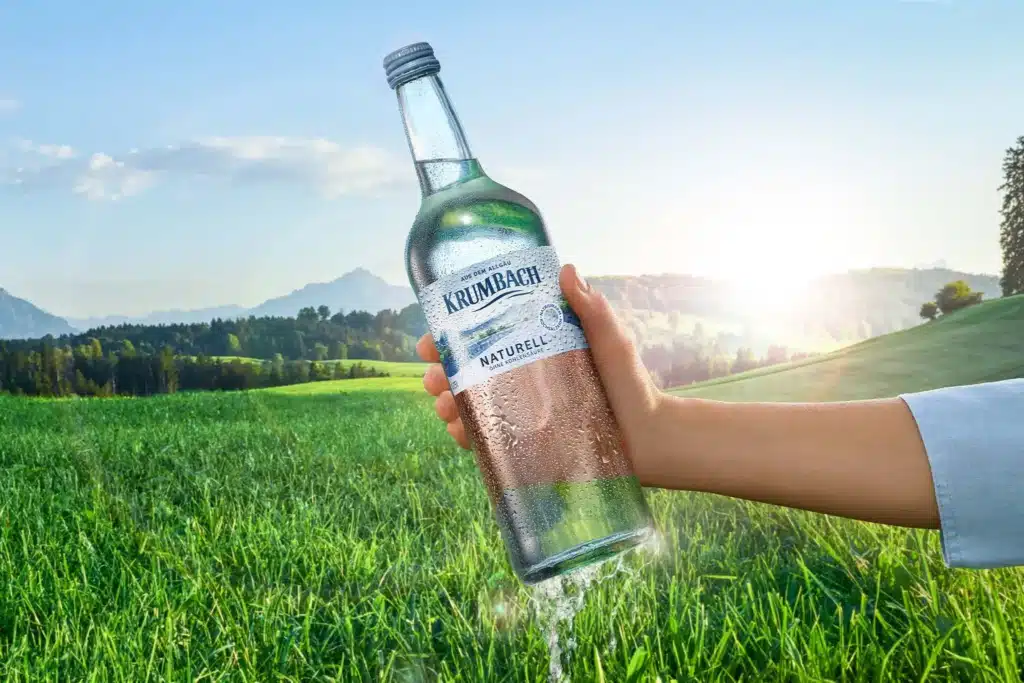 Hand hält gekühlte Krumbach Naturell Genussflasche vor grüner Allgäuer Landschaft mit Bergen und Sonne
