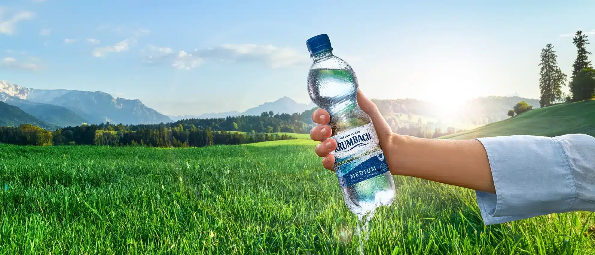 Frau hält Krumbach Mineralwasserflasche vor idyllischer Graslandschaft