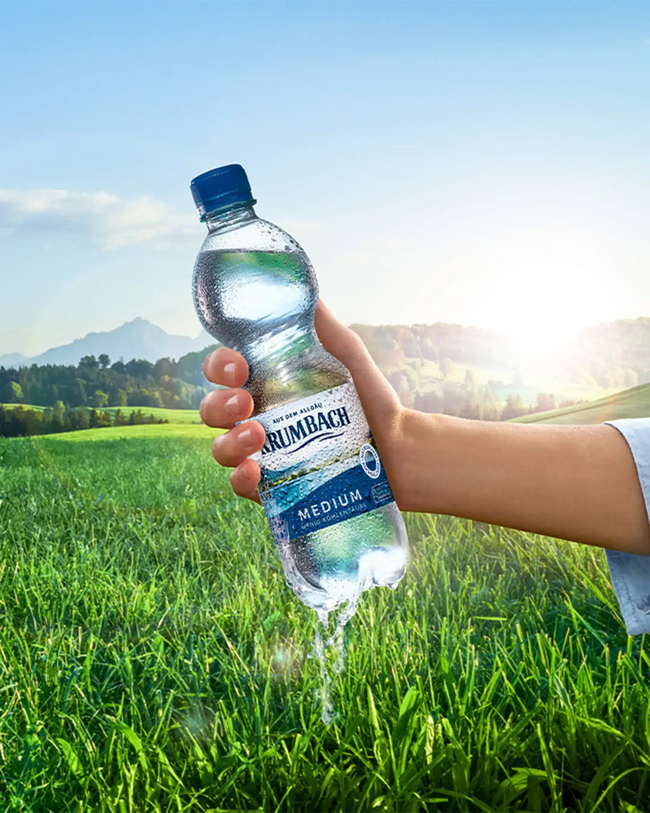 Frau hält Krumbach Mineralwasserflasche vor idyllischer Graslandschaft