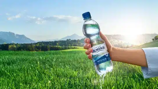 Frau hält Krumbach Mineralwasserflasche vor idyllischer Graslandschaft