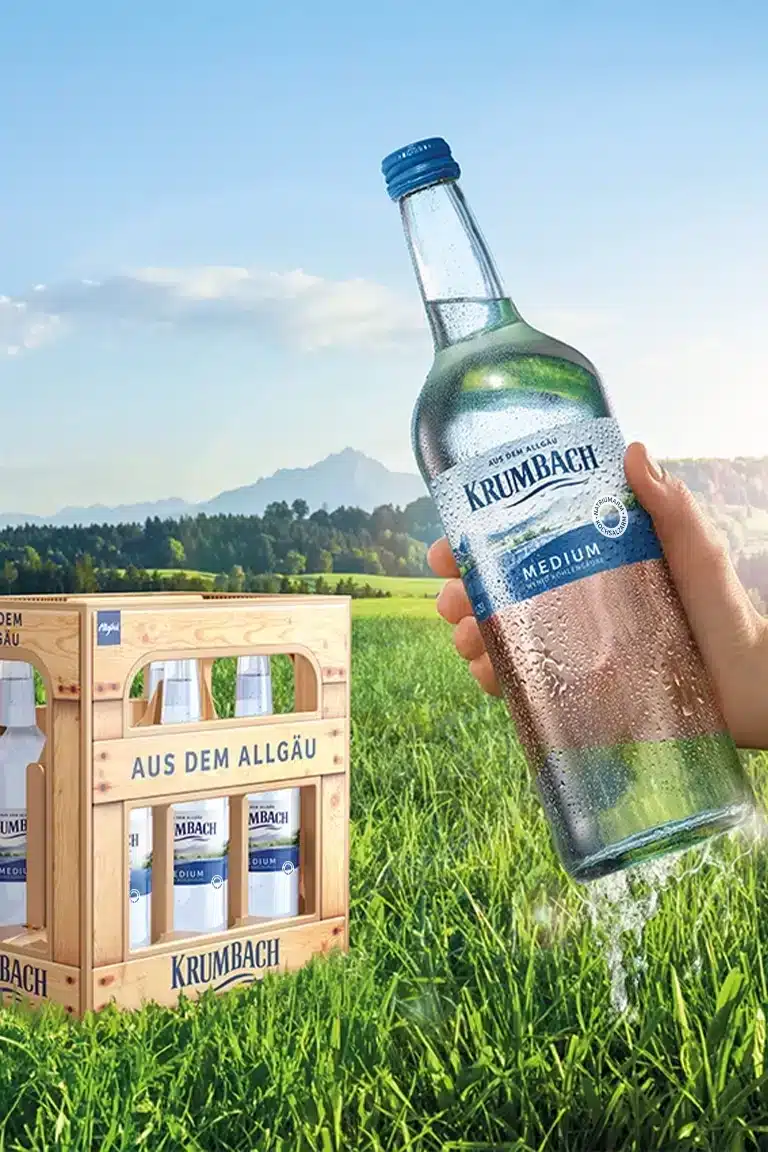Hand hält Krumbach Mineralwasser (Medium) vor Allgäu-Kulisse, daneben 6er-Kasten