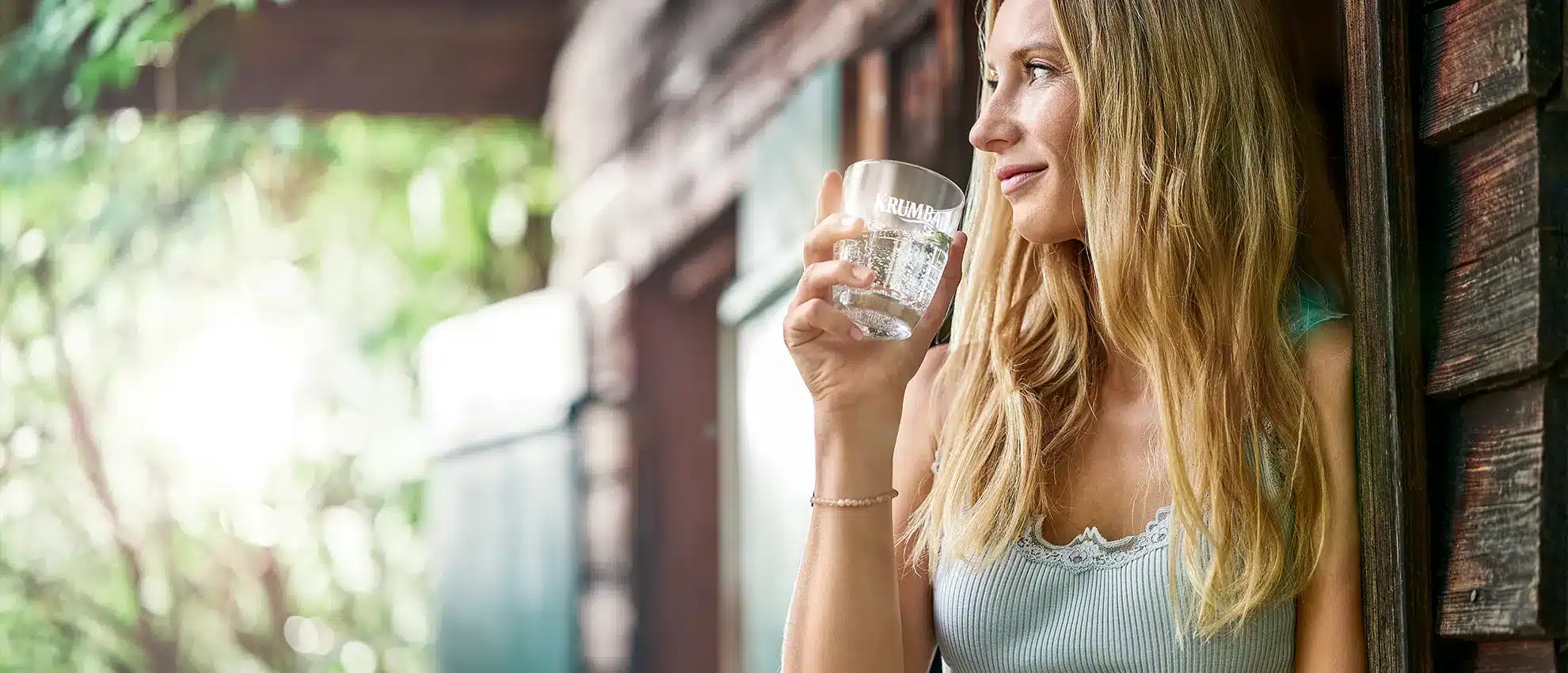 Blonde Frau hält ein Glas Krumbach Mineralwasser und blickt entspannt aus einem geöffneten Fenster ins Grüne