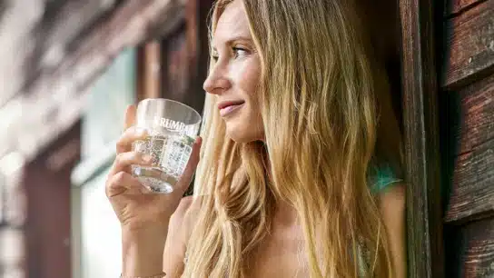 Blonde Frau hält ein Glas Krumbach Mineralwasser und blickt entspannt aus einem geöffneten Fenster ins Grüne