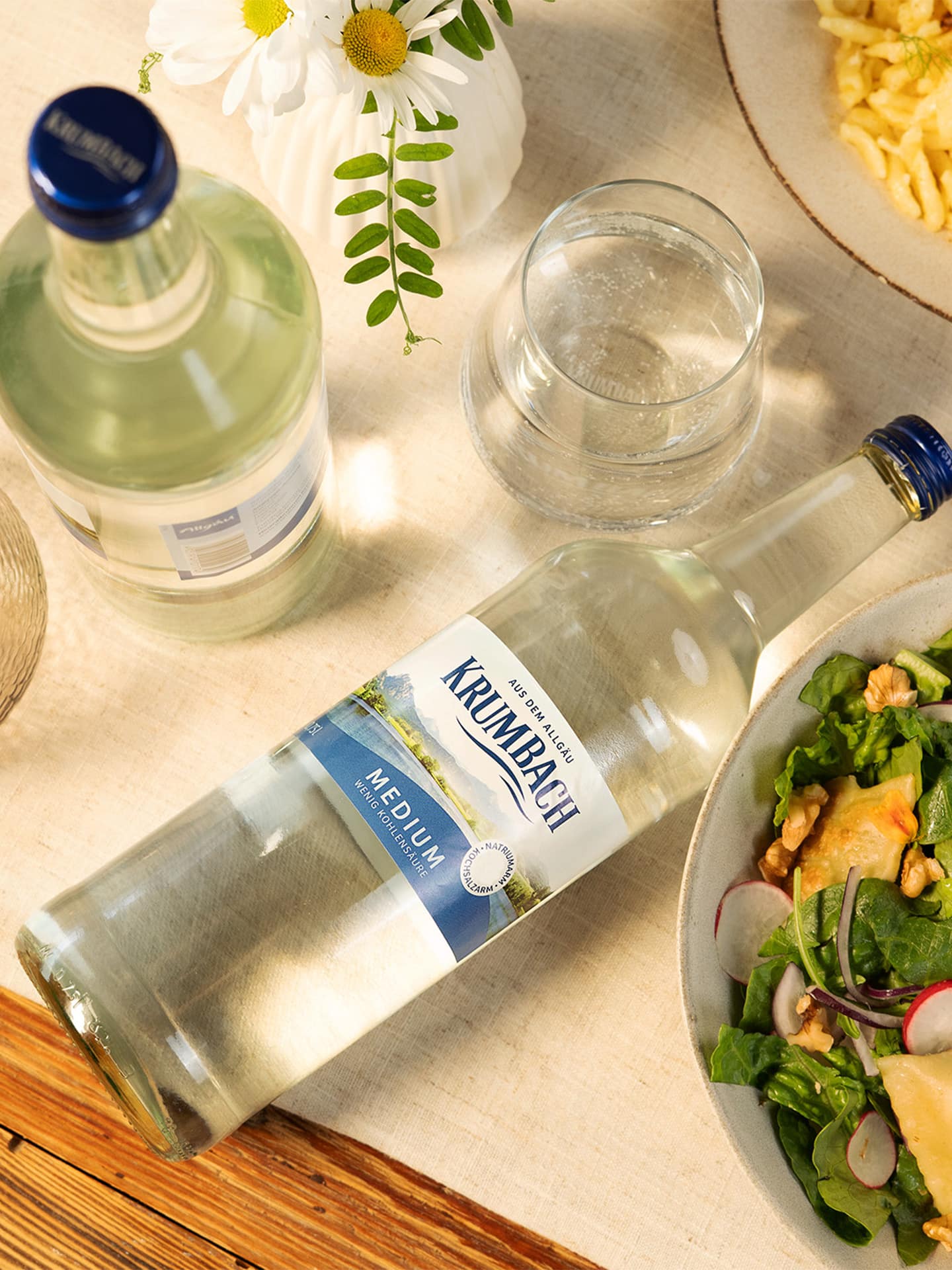 Krumbach Medium Mineralwasser auf gedecktem Tisch mit Glas, Blumen und Speisen in sonniger Atmosphäre